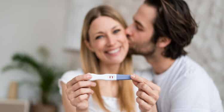 Happy young couple showing positive pregnancy test and kissing at home, selective focus